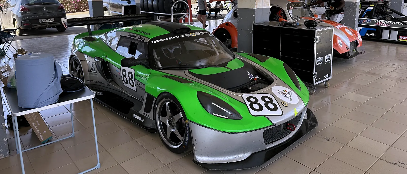 Zelený Exige č. 88 v garáži okruhu, za ním vozy soupeřů, Circuit de Baobab