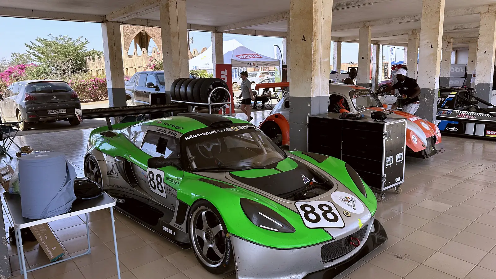 Exige 88 v boxu Circuit de Baobab, za ním černý vůz, sloupy a bugenvilea, Senegal