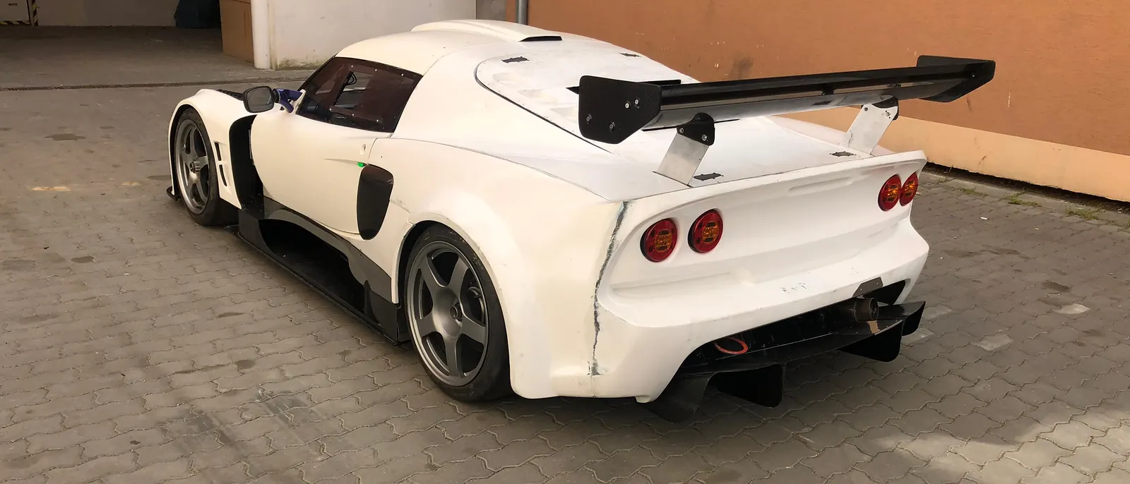 Bílý Lotus Exige v gelcoatu zezadu na dvoře dílny, křídlo a difuzor